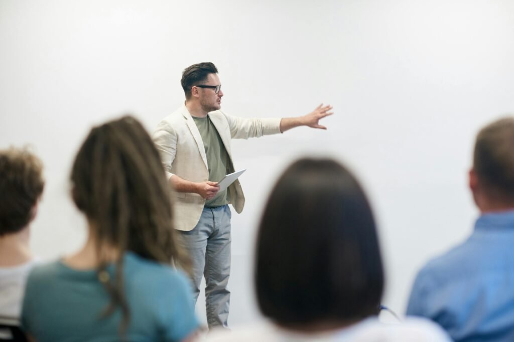 Un homme en veste beige et t-shirt vert, tenant une tablette et donnant une présentation à un groupe de personnes assises. Le but de l'intervention, est de sensibiliser les collaborateurs à des process de management responsable.